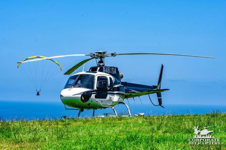 Helicopter on a grassy field, overlooking the ocean. A paraglider flies in the background.