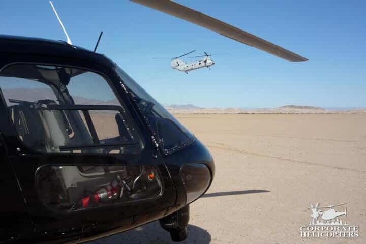 A military helicopter flies in the background of a civilian helicopter on the ground